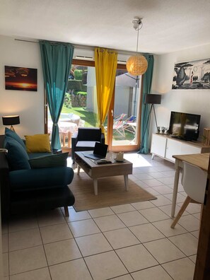 A living room with a sofa, coffee table, and a TV. There is a window with curtains, a lamp, and a painting on the wall. - Apartment 'Cosy Entre Lac Et Montagnes' with Private Terrace (Thônes)