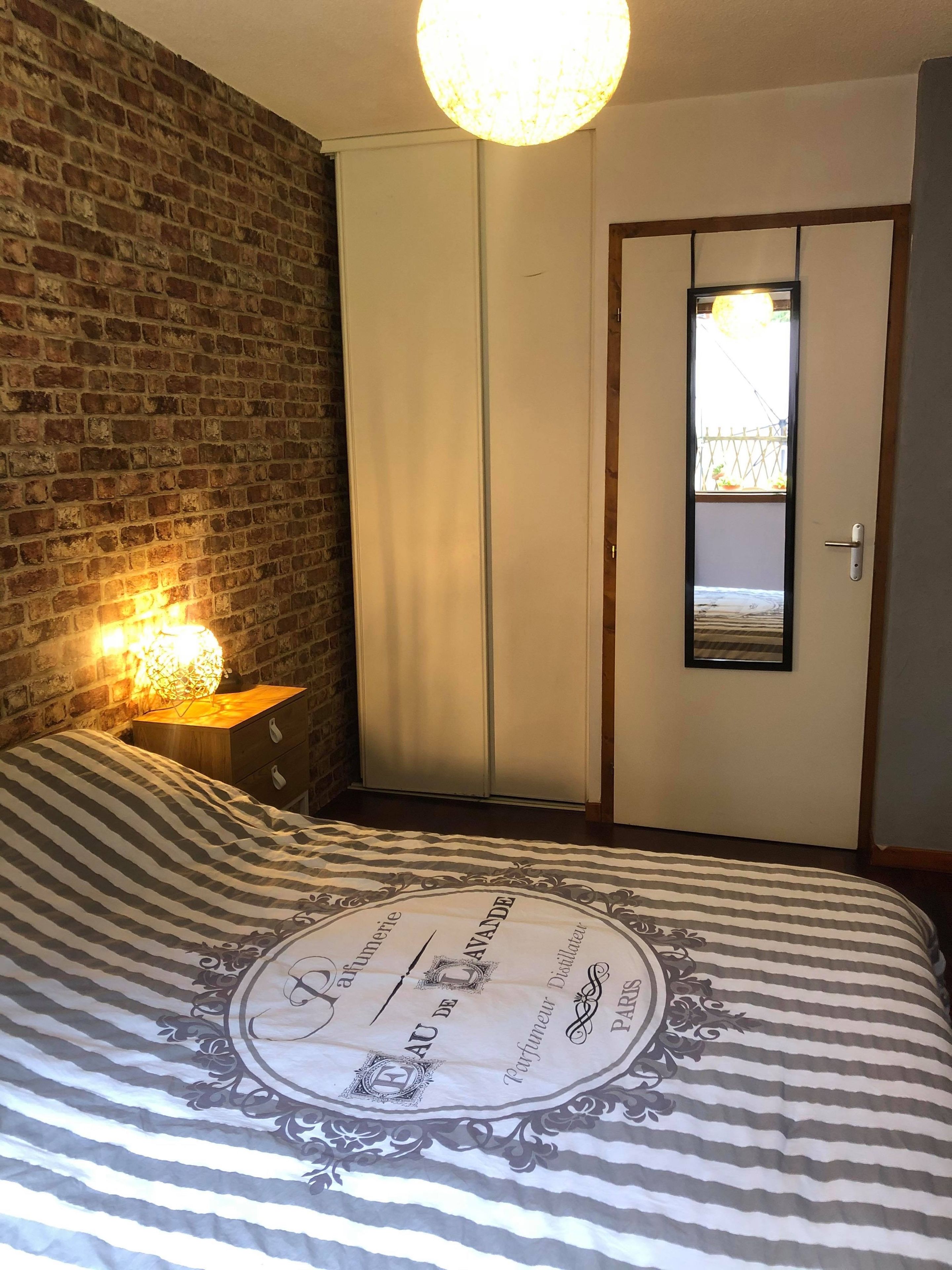 Une chambre avec une couvre-lit à rayures, un mur en briques, une table de chevet en bois avec une lampe et une armoire avec un miroir.