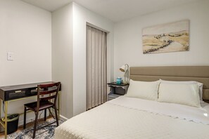 A hotel room with a bed, a desk, a chair, a nightstand, a lamp, and a painting on the wall.