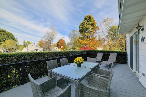 An outdoor patio with a glass table and wicker chairs, surrounded by a hedge and trees with autumn foliage.