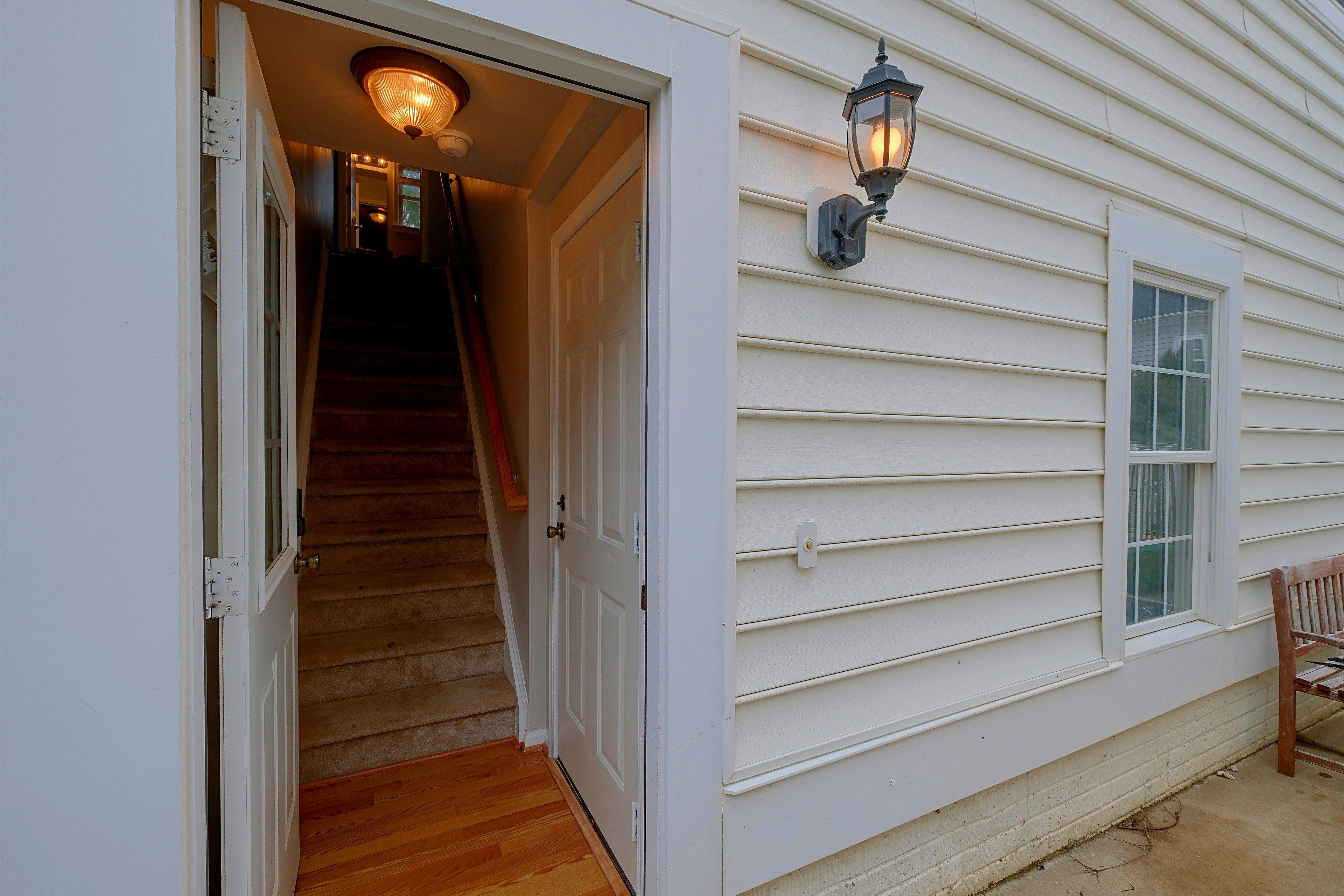A white exterior wall with a window and a door, a wooden staircase, and a wall-mounted light fixture.