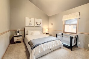 A bedroom with a bed, bedside table, lamp, artwork, window, and a crib.