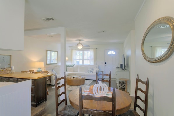 A dining area with a wooden table, chairs, and a round ottoman. There is a ceiling fan, a window with curtains, and a white door in the background.