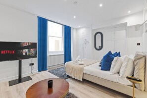 Una sala de estar moderna con un televisor de pantalla plana, una mesa de centro redonda, un sofá con cojines azules y blancos, y un ventanal con cortinas azules.