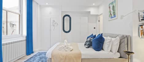 A modern bedroom with a bed, a bedside table, a lamp, a window with curtains, and a wall with a framed picture.