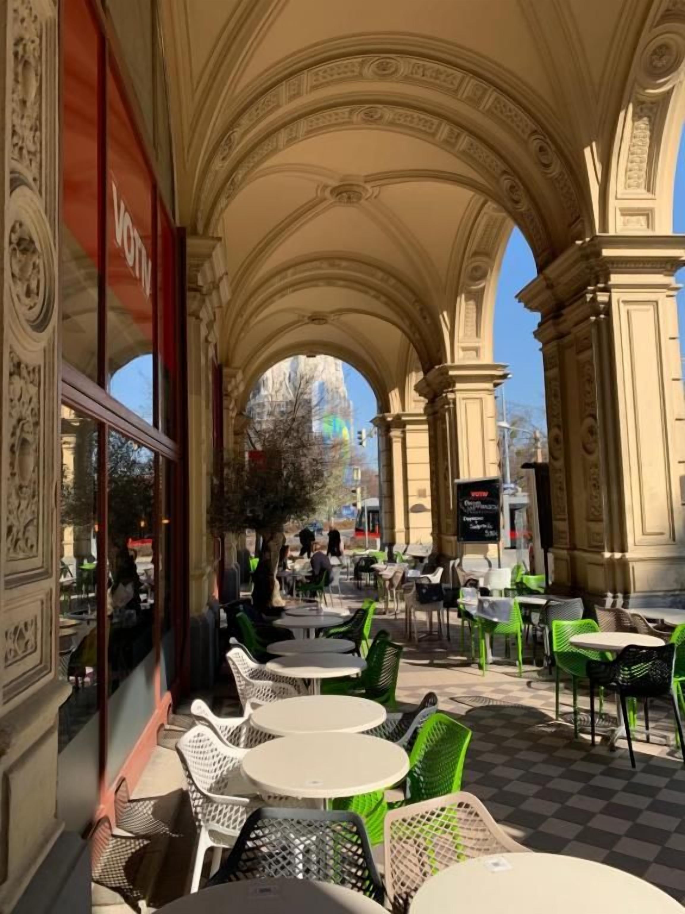 An outdoor cafe with ornate arches, round tables, and green chairs.