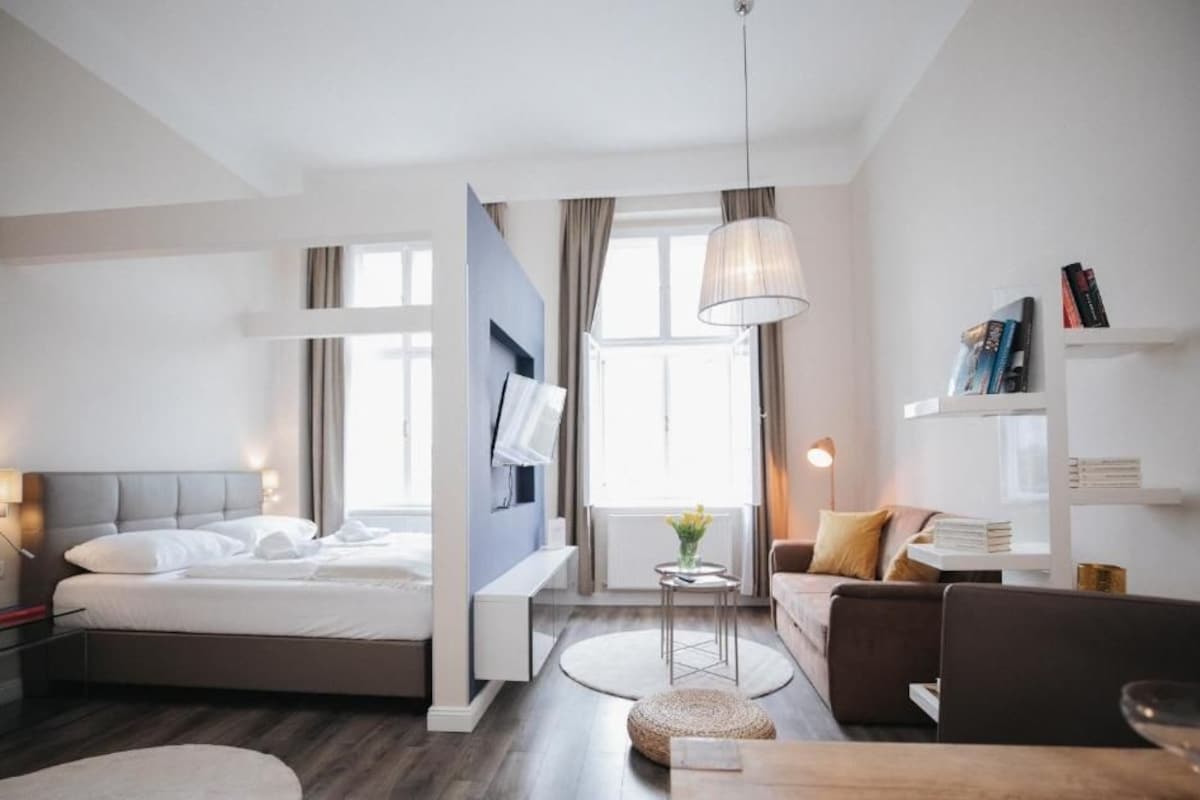 A modern hotel room with a bed, bedside table, lamp, and a seating area with a sofa and a small table.