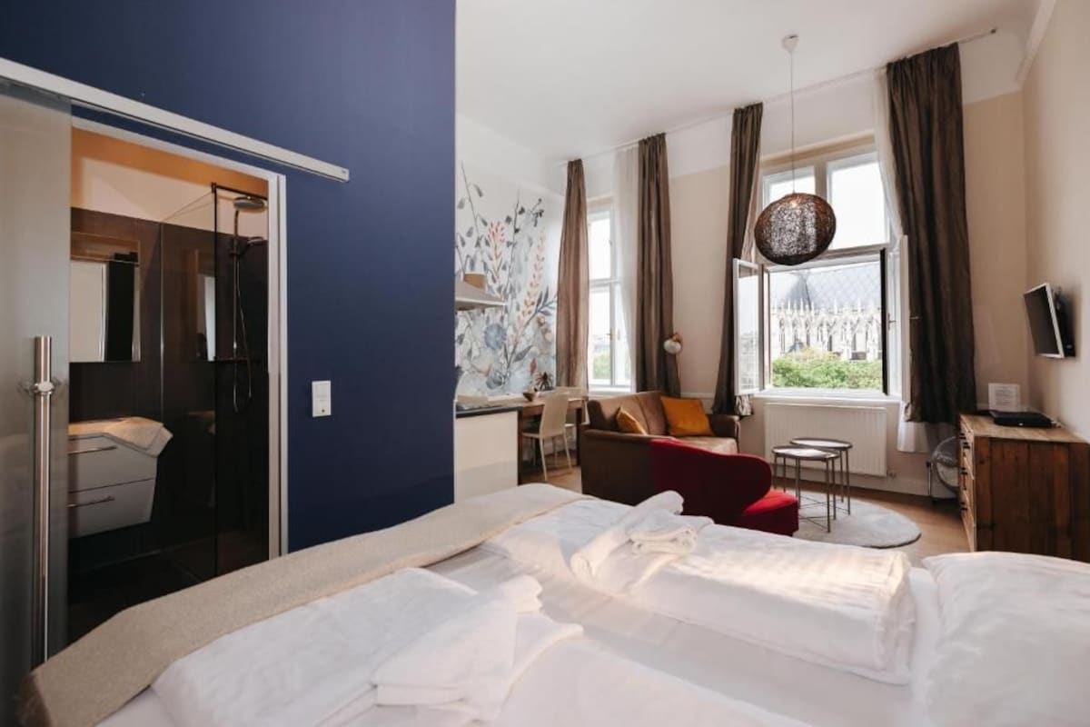 A hotel room with a bed, a desk, a chair, a red sofa, and a window with curtains.