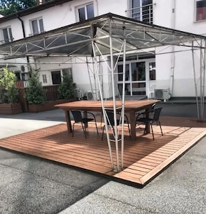 An outdoor patio with a wooden table and chairs, a transparent roof structure, and a white building in the background. - Astoria Apart Hotel (Skåne län)
