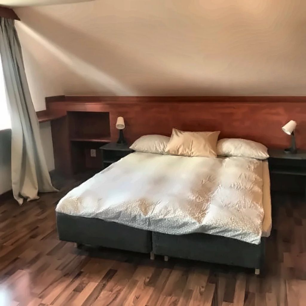 Un dormitorio con piso de madera, una cama con colcha blanca y dorada, dos mesitas de noche con lámparas y un cabecero de madera.