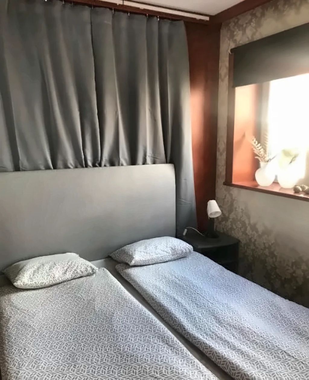A room with two beds, a nightstand, a lamp, and a window with curtains.