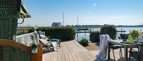 A wooden deck with lounge chairs, a table with a vase of flowers, and a view of a marina with boats.