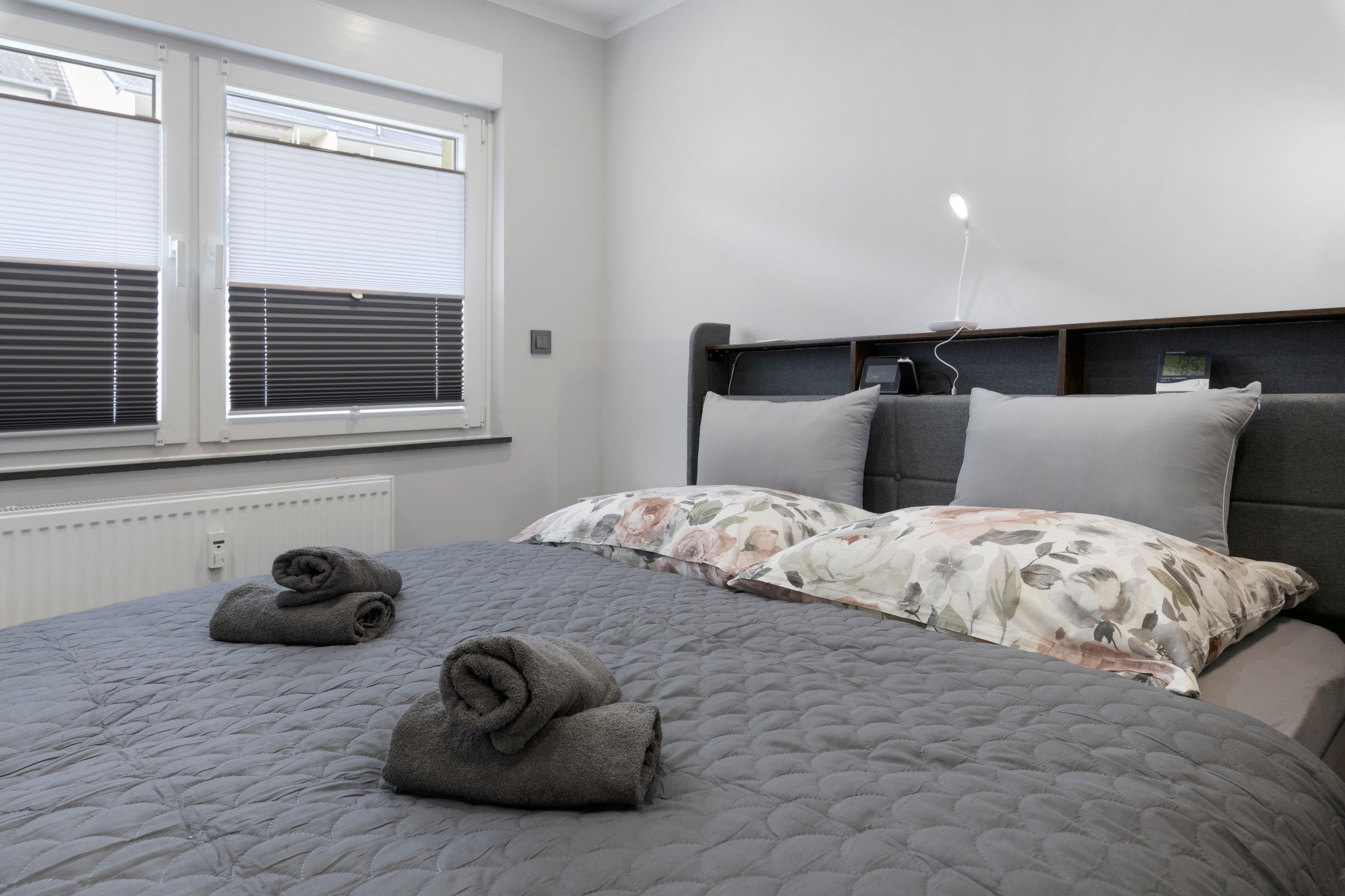 Ein Schlafzimmer mit einem Bett, zwei gerollten Handtüchern, einem Fenster mit Jalousien, einem Thermostat und einem Nachttisch mit einer Lampe.
