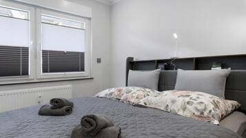 Ein Schlafzimmer mit einem Bett, zwei gerollten Handtüchern, einem Fenster mit Jalousien, einem Thermostat und einem Nachttisch mit einer Lampe.