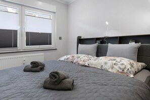 Ein Schlafzimmer mit einem Bett, zwei gerollten Handtüchern, einem Fenster mit Jalousien, einem Thermostat und einem Nachttisch mit einer Lampe.