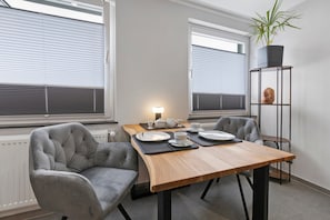 Ein moderner Essbereich mit einem Holztisch, zwei grauen Polsterstühlen und einer schwarzmetallenen Kommode mit einer Pflanze.
