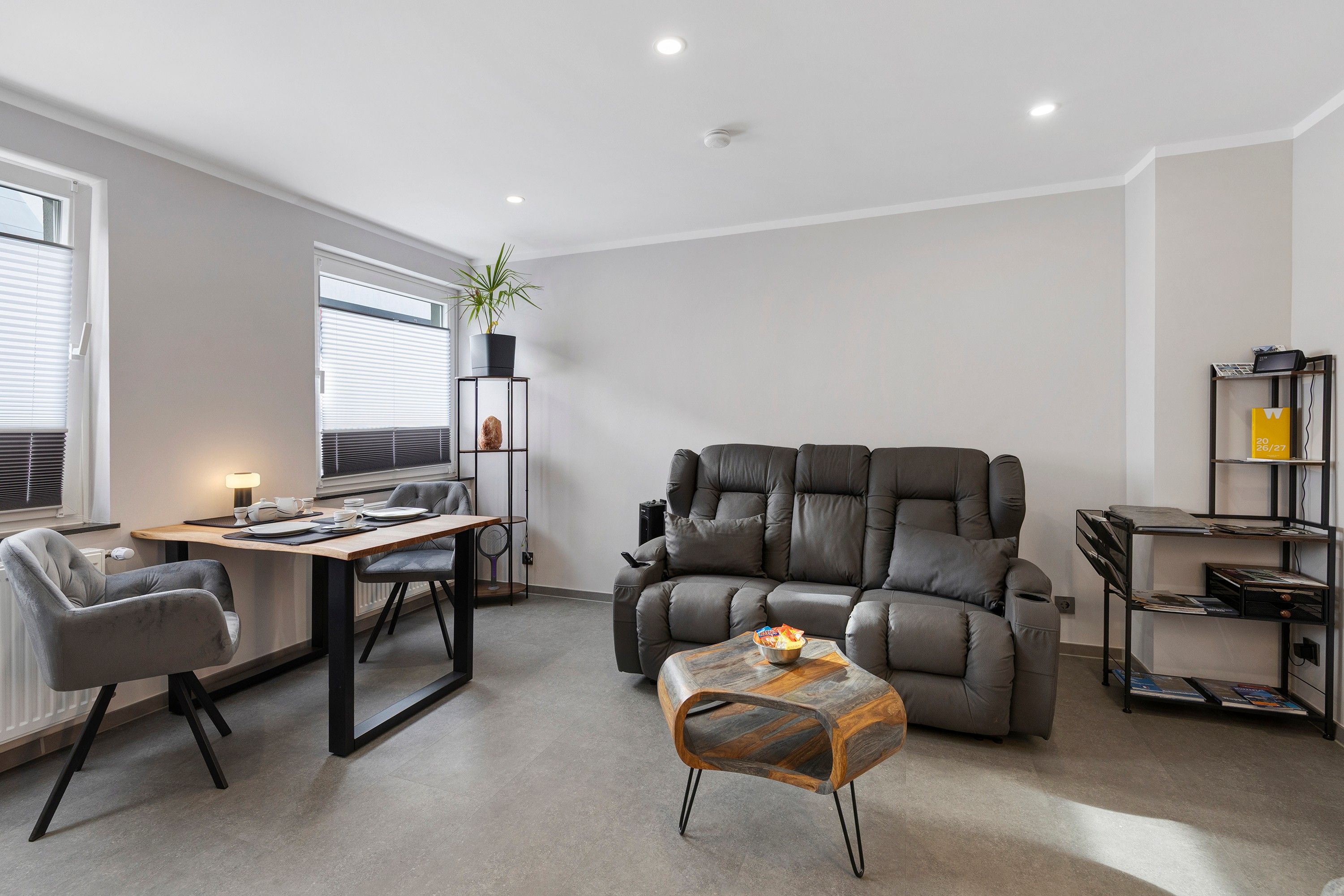 Ein modernes Wohnzimmer mit einer Ledersofa, einem Esstisch mit Stühlen, einem kleinen Couchtisch und einem Bücherregal.