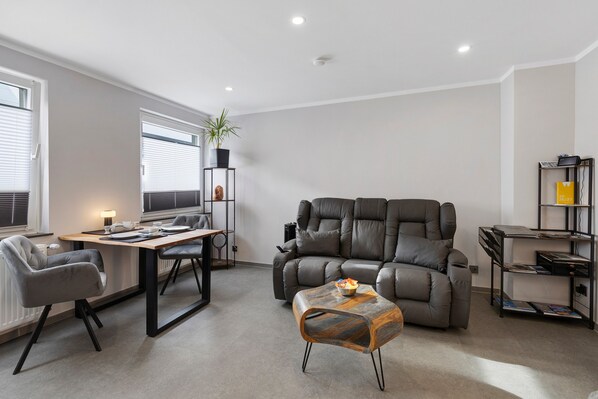 Ein modernes Wohnzimmer mit einer Ledersofa, einem Esstisch mit Stühlen, einem kleinen Couchtisch und einem Bücherregal.