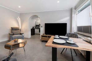 A modern living room with a sofa, a dining table set for two, a flat-screen TV, and a kitchen in the background.