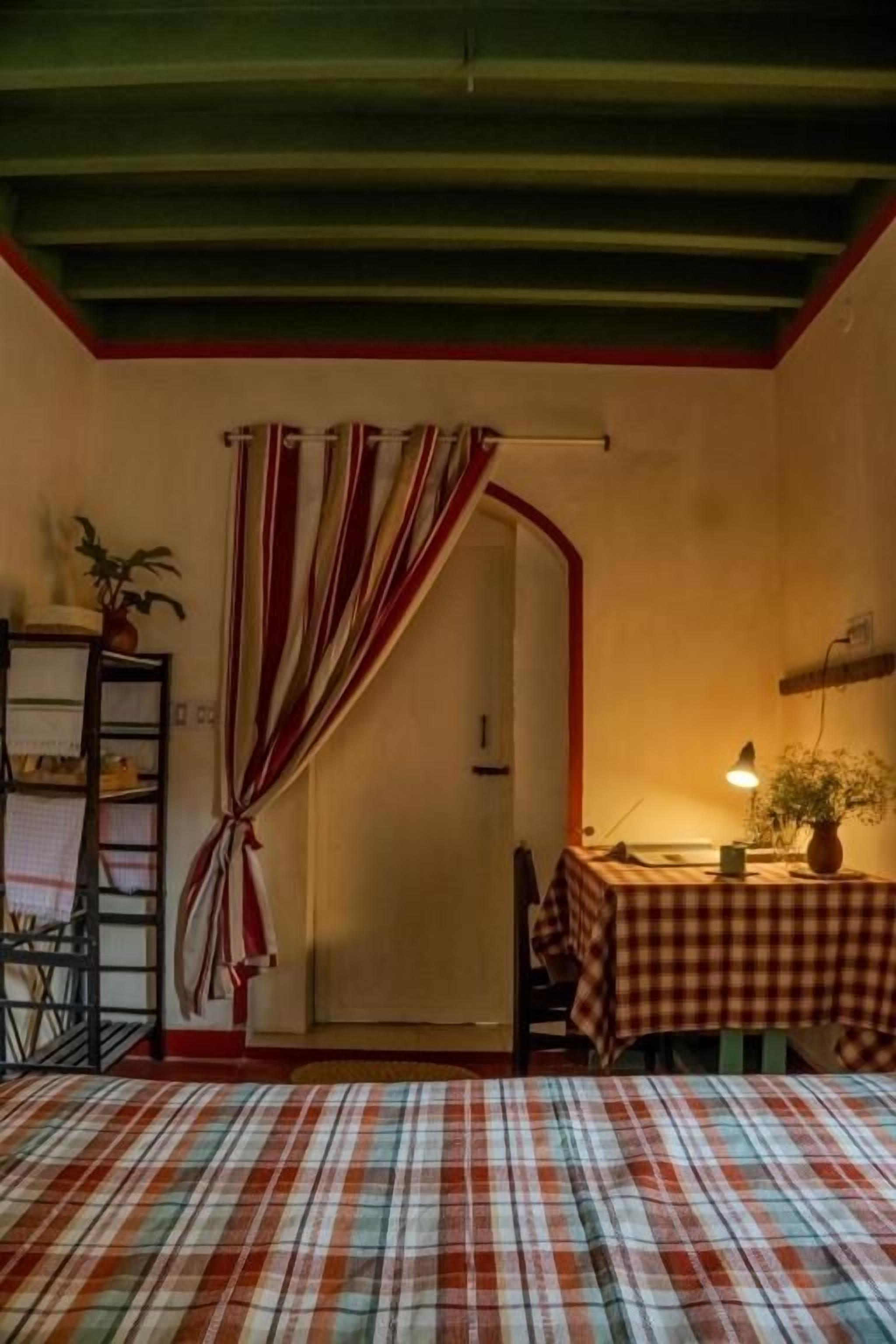 A room with a bed, a table with a lamp, a chair, a shelf with a plant, and a curtain.