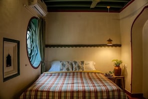 Un dormitorio con cama, ventana con cortinas, aire acondicionado instalado en la pared, una mesita con una planta y un aplique en la pared.