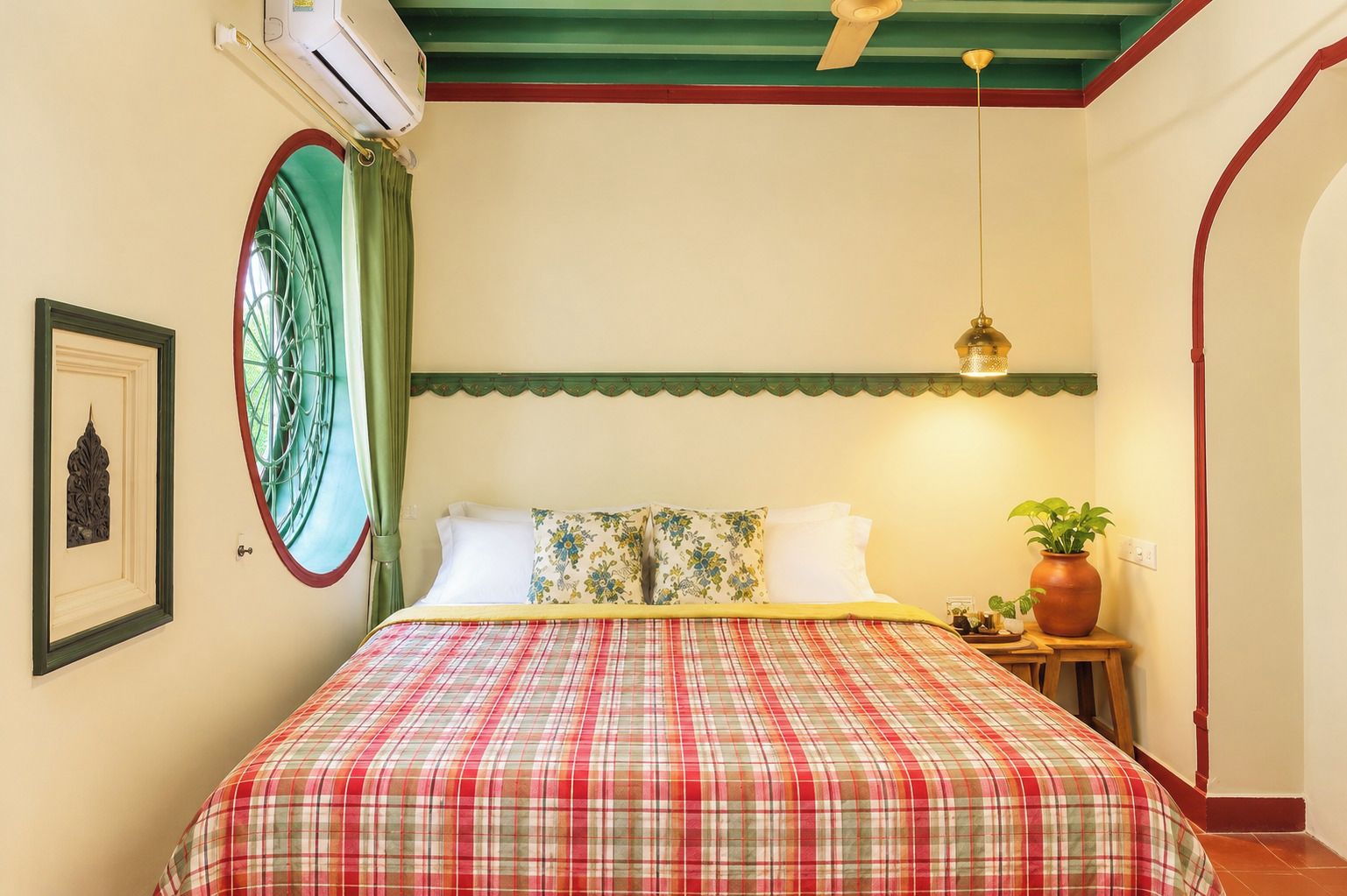 A bedroom with a bed, a window with curtains, a ceiling fan, a wall-mounted air conditioner, and a bedside table with a plant.