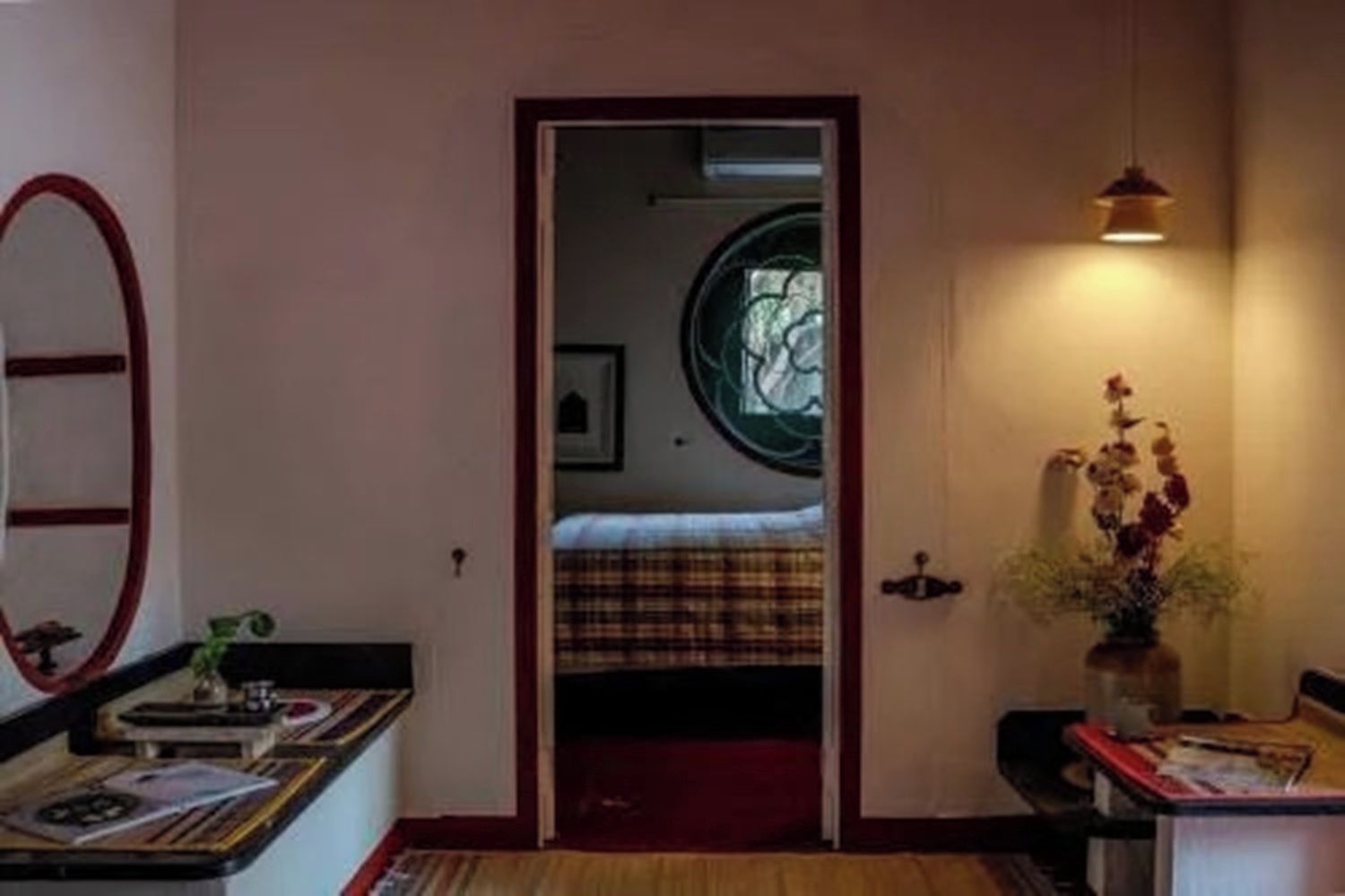 A room with a bed, a mirror, a vase with flowers, and a window with a circular design.