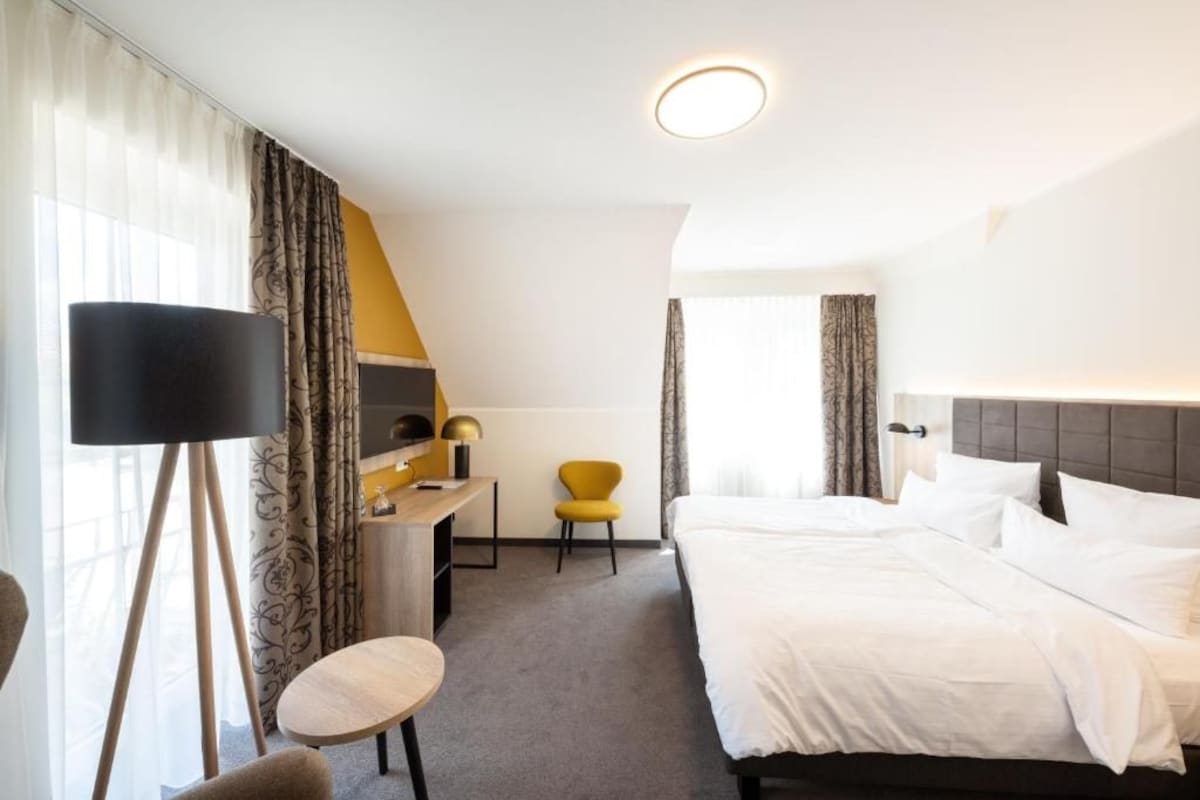 Une chambre d’hôtel avec un lit, un bureau doté d’une lampe, une chaise et une grande fenêtre avec des rideaux.