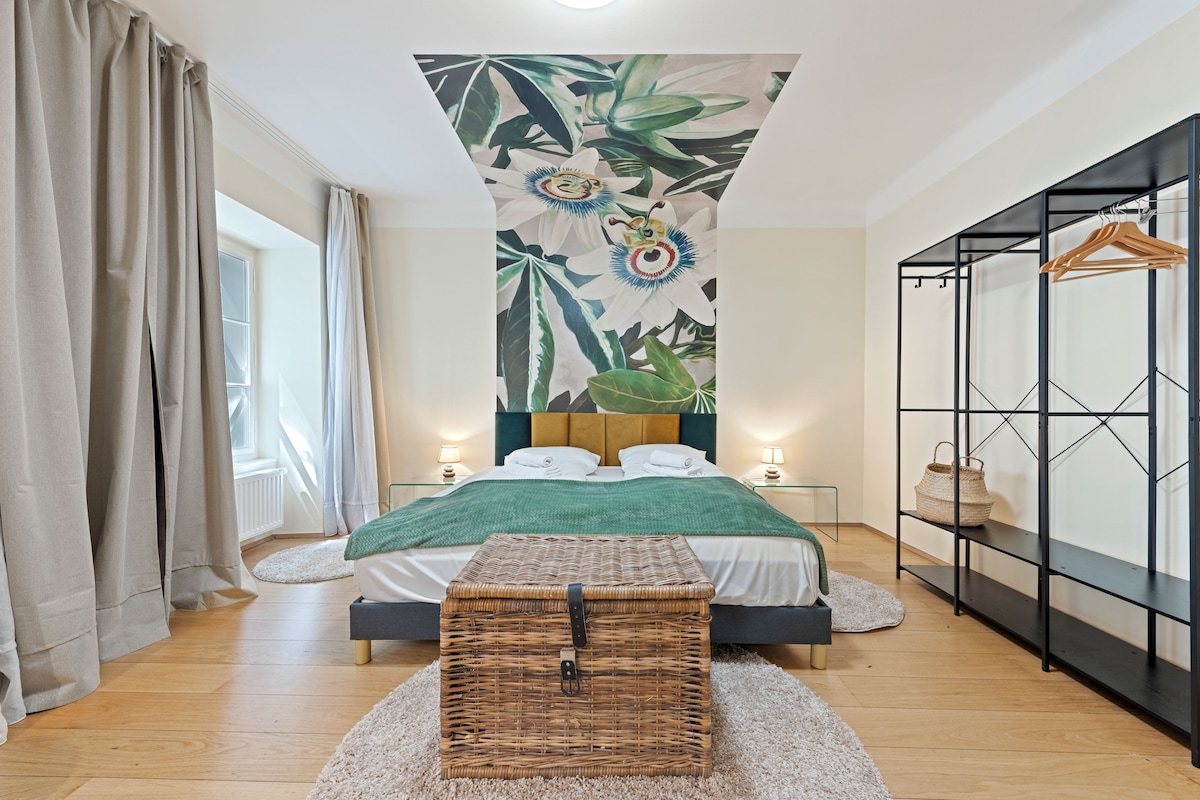Een hotelkamer met een bed, een groot raam met gordijnen, een wandplank en een hoofdbord met een tropisch dessin.