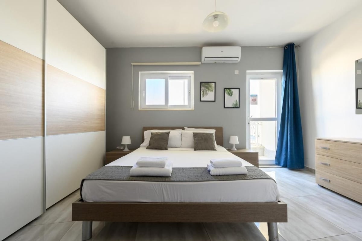 Ein modernes Schlafzimmer mit einem großen Bett, Nachttischen, einer Klimaanlage und Blick ins Freie durch ein Fenster.