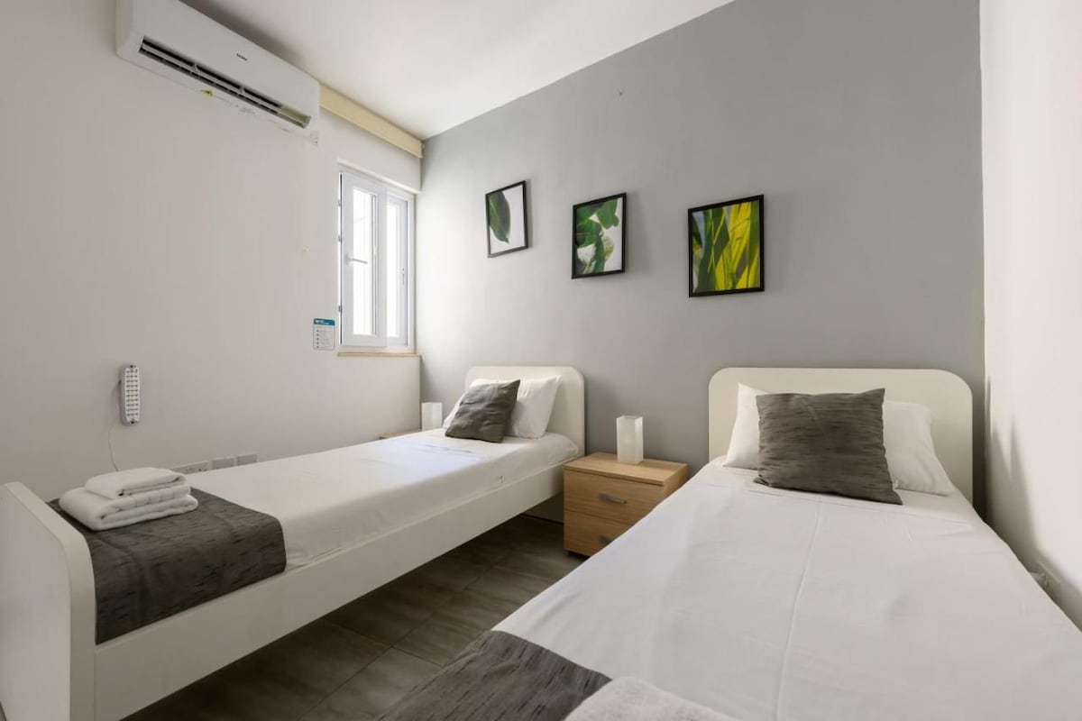 Ein Doppelzimmer mit zwei Einzelbetten, einem Nachttisch mit Lampe, einem Fenster und Wandbildern.