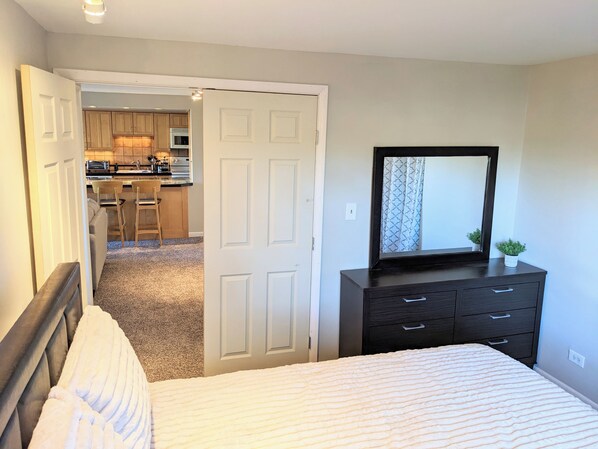 A bedroom with a bed, a dresser with a mirror, and an open door leading to a kitchen area. - 30 Day+ Quiet, 2 BR/1 BA Condo in Congress Park (Denver)