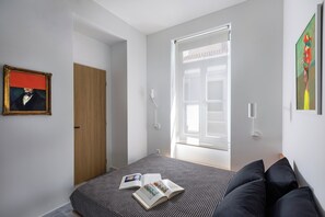 A bedroom with a bed, a window, a door, and a painting on the wall. - Art Space Residencies Α1 (Athina)