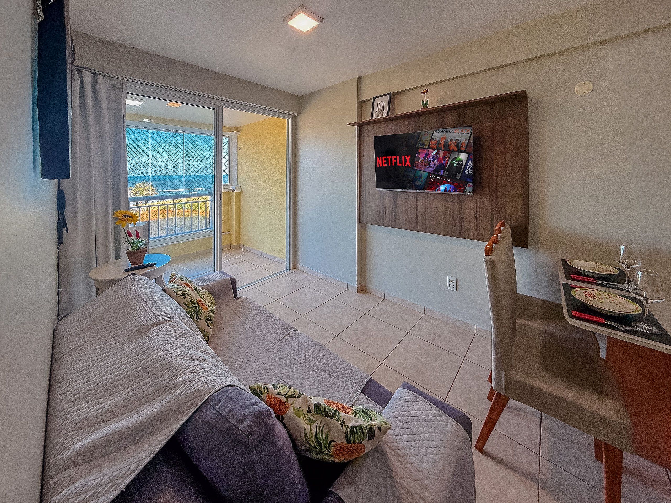 Una sala de estar con sofá, televisión instalada en la pared, balcón con vistas y zona de comedor con mesa.