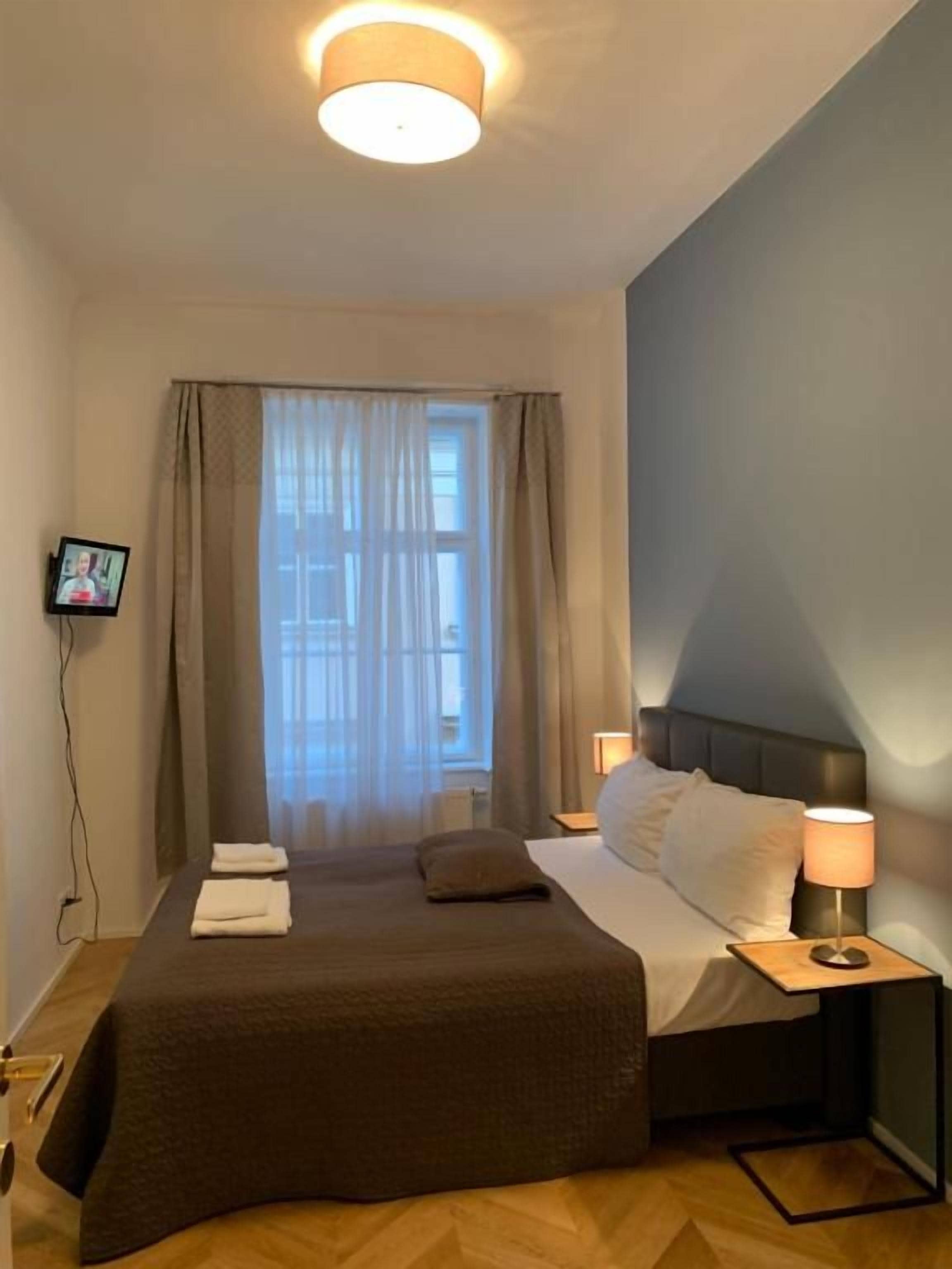 A hotel room with a bed, a TV mounted on the wall, a window with curtains, and a bedside table with a lamp.