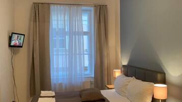 A hotel room with a bed, a TV mounted on the wall, a window with curtains, and a bedside table with a lamp.
