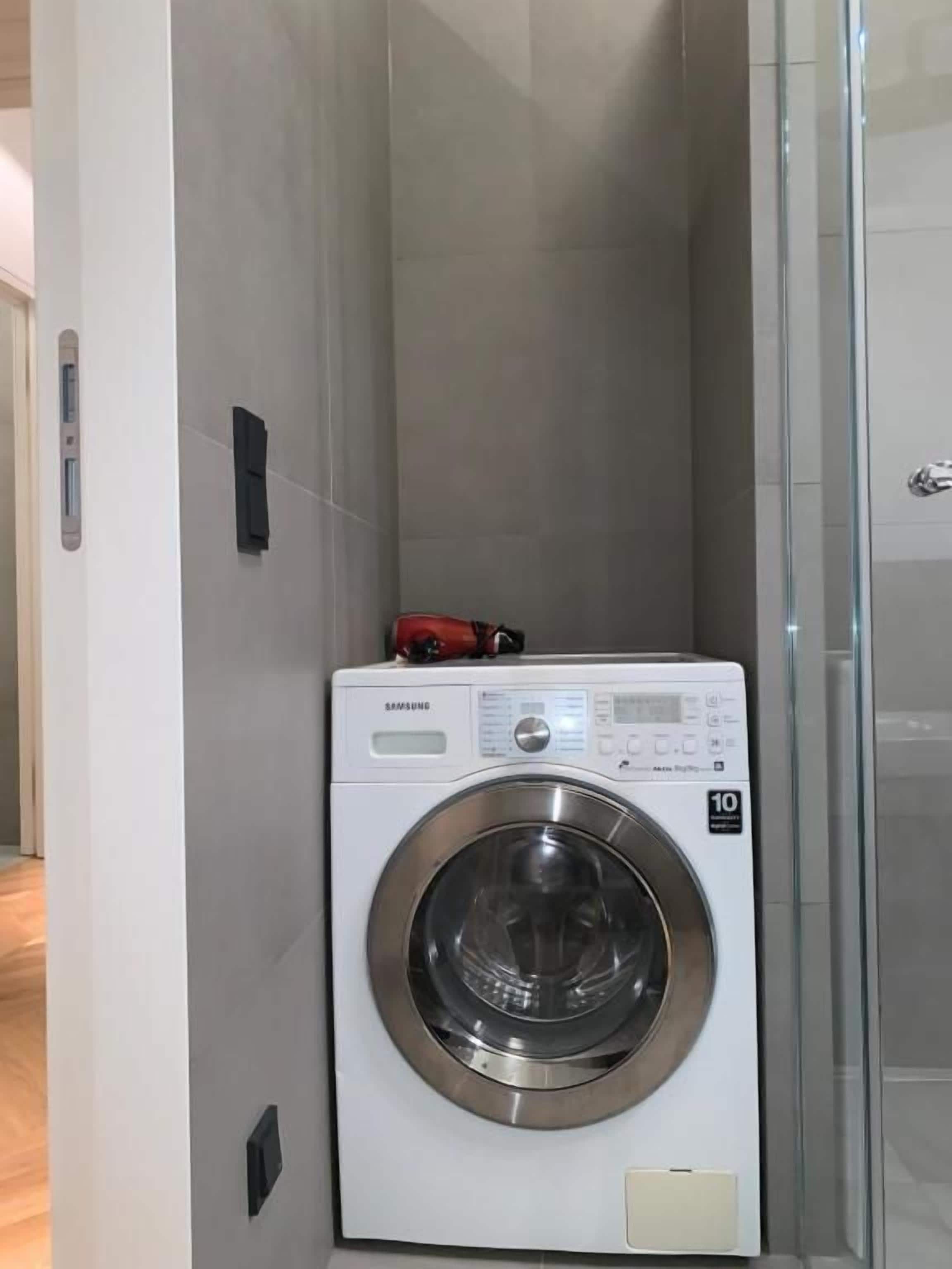 Una lavadora compacta de carga frontal, blanca, con panel de control digital, ubicada en un lavadero estrecho y con azulejos contiguo a un box de ducha de vidrio.