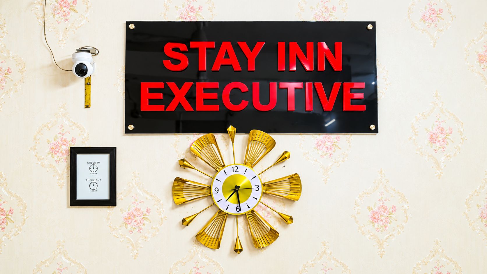 A wall with a sign that reads 'STAY INN EXECUTIVE,' a security camera, a framed clock, and a decorative wall clock.