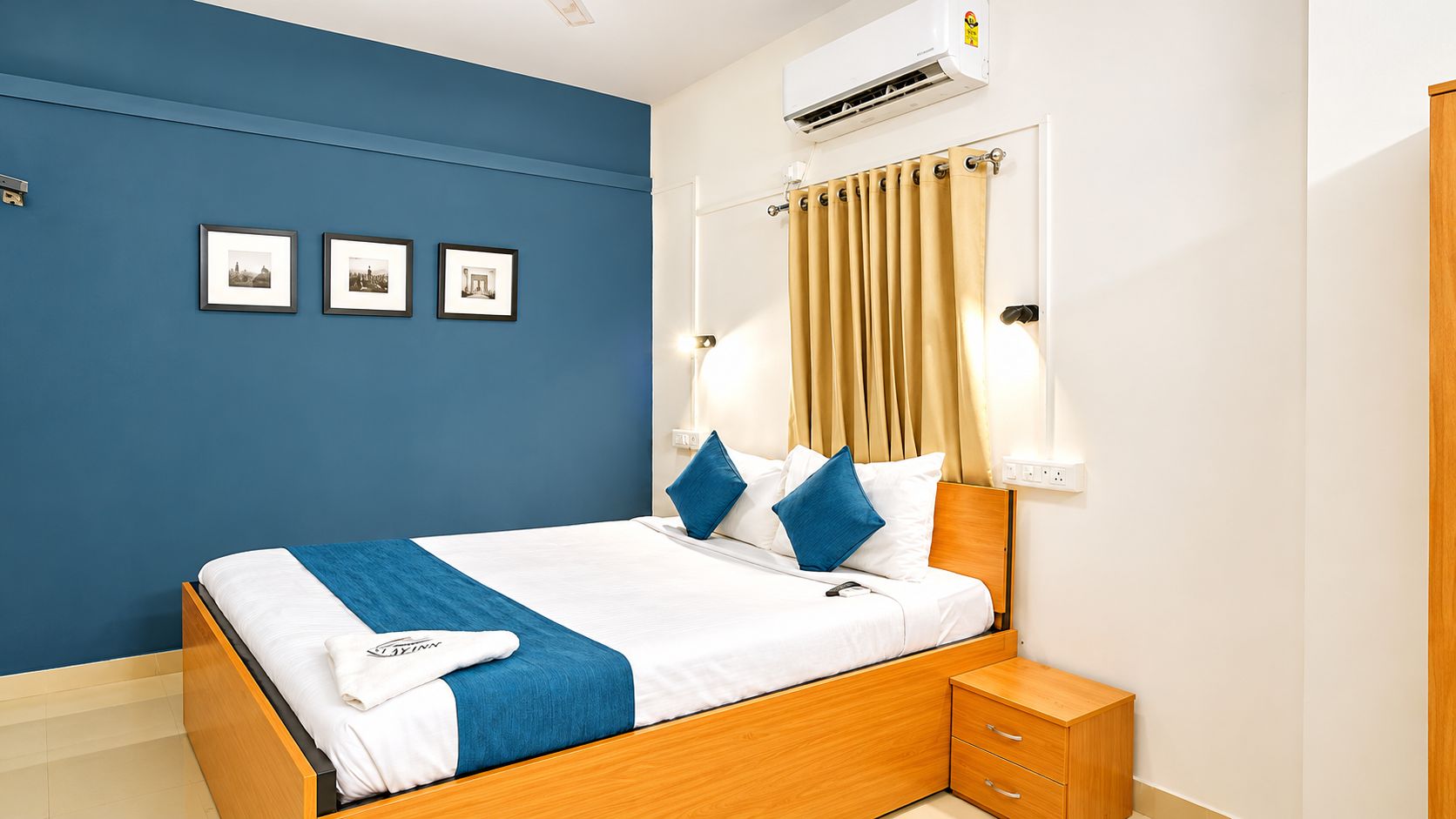 A hotel room with a bed, a nightstand, a wall-mounted air conditioner, and three framed pictures on the wall.