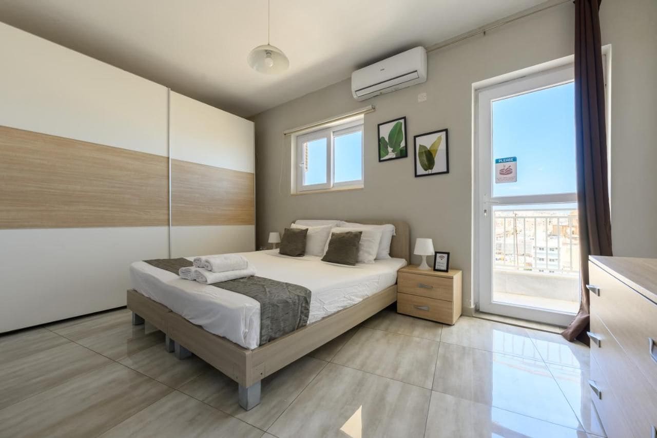 Ένα σύγχρονο υπνοδωμάτιο με κρεβάτι, κομοδίνα, μια ντουλάπα και ένα μπαλκόνι με θέα.