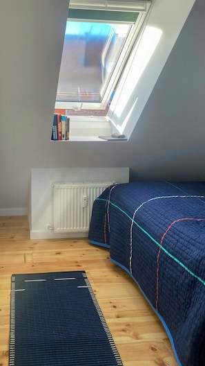 Ein kleines, gut beleuchtetes Schlafzimmer im Dachgeschoss mit einem Oberlicht, einem Heizkörper, einem Bett mit Patchworkdecke und einem Teppich auf dem Boden.