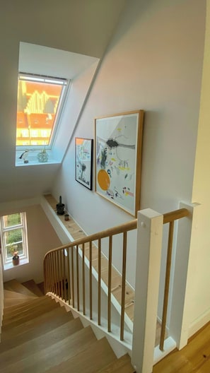 Eine Treppe mit hölzernen Geländern führt zu einer oberen Etage, ein Oberlicht und zwei gerahmte Kunstwerke an der Wand sind zu sehen.