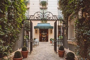 Un étroit sentier pavé de pierres menant à l’entrée d’un hôtel, avec un portail en fer forgé, des plantes en pot et des sièges installés à l’extérieur.