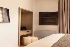 Une chambre d’hôtel comprenant un lit, une armoire en bois, un petit tabouret, un téléviseur à écran plat et une fenêtre avec des stores.