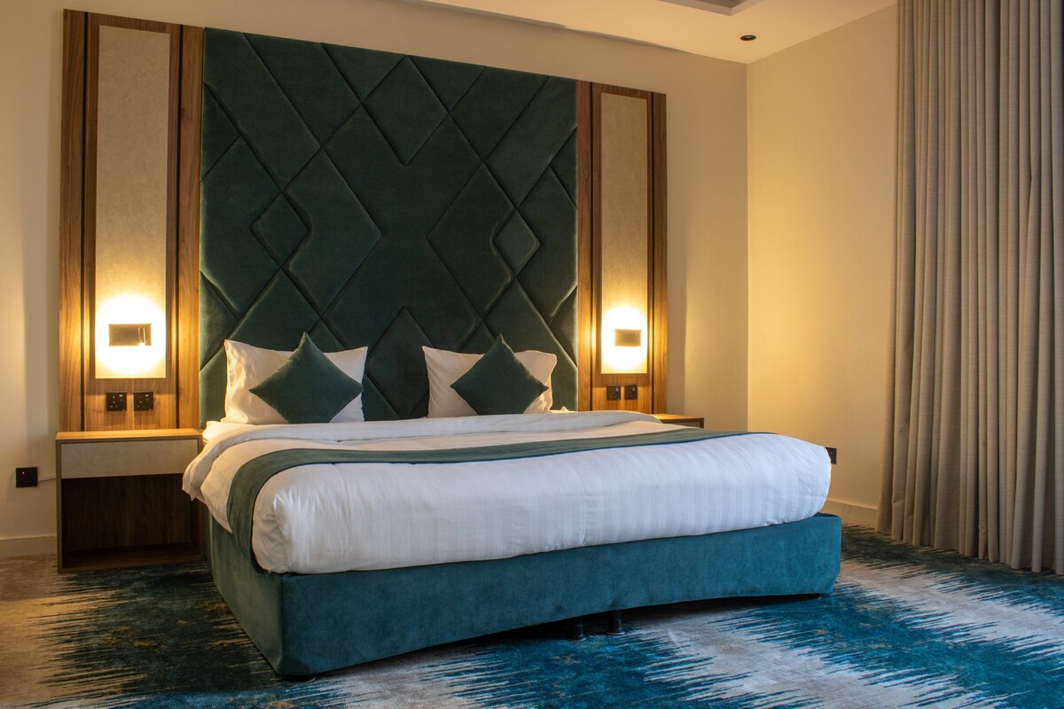 A hotel room with a large bed, a tufted headboard, two bedside lamps, and a blue patterned carpet.