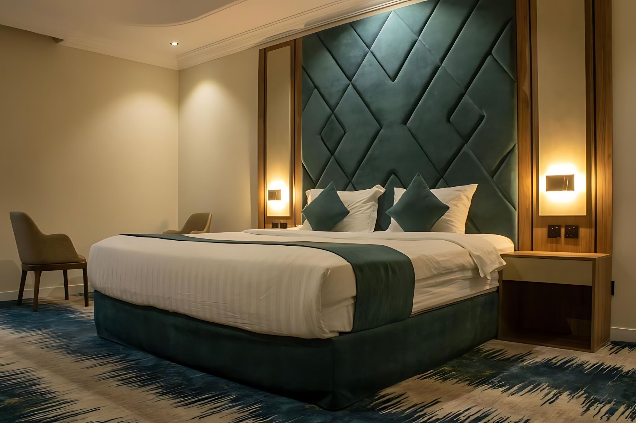 Habitación de hotel con una cama grande, dos mesitas de noche, una silla y un cabecero moderno.