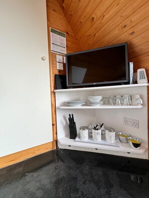 A kitchenette with a flat-screen TV, white shelves with plates, glasses, and utensils, and a wooden ceiling.
