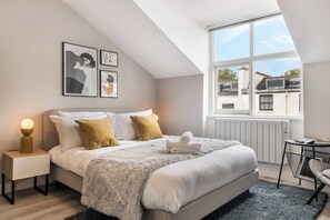 Une chambre moderne avec un grand lit, une table de chevet avec une lampe, une fenêtre donnant sur des immeubles et des œuvres d’art accrochées au mur.
