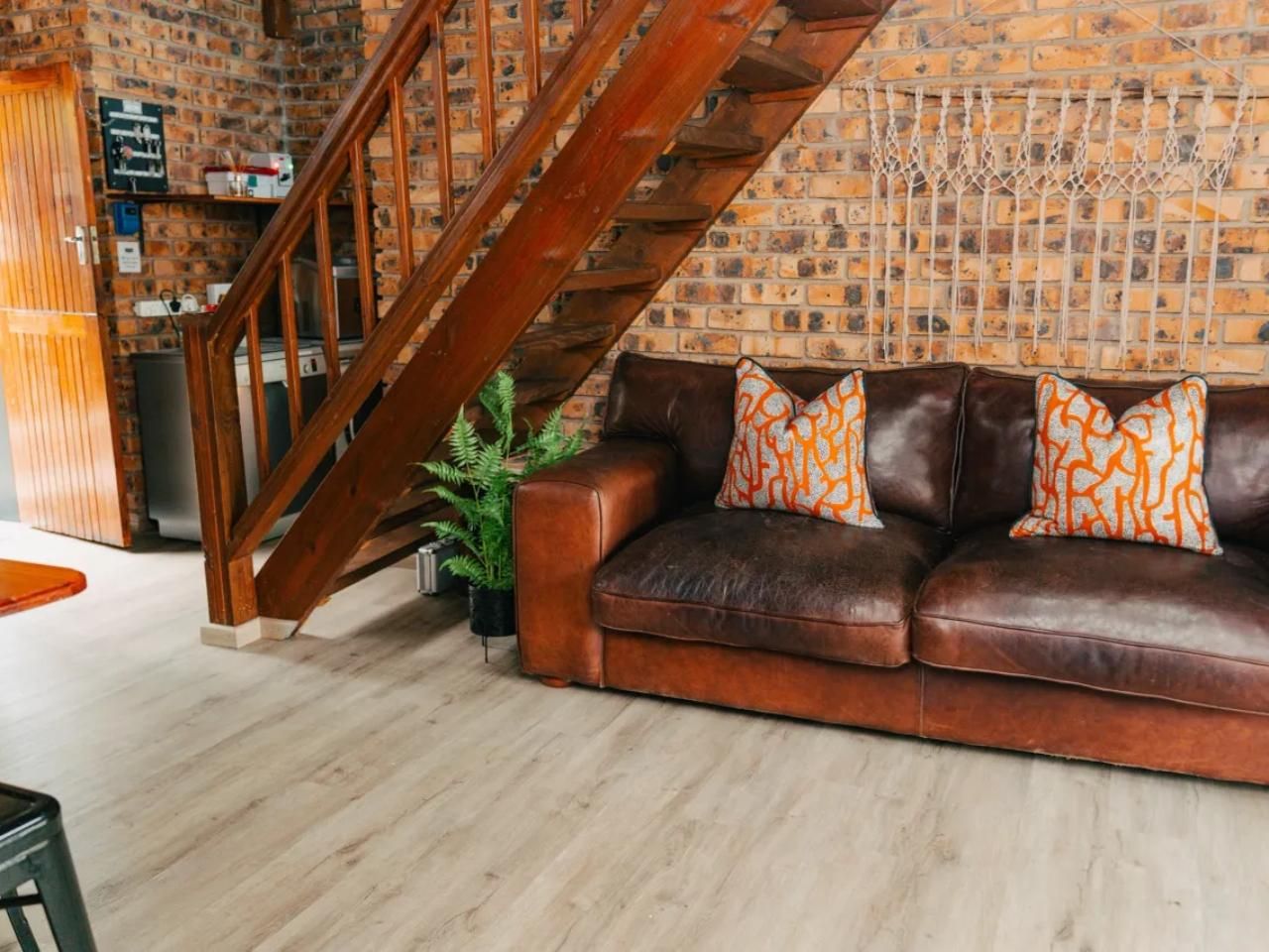 A living room with a brown leather sofa, orange patterned cushions, a wooden staircase, and a brick wall.