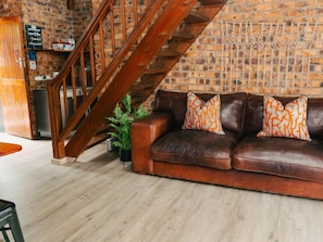 Una sala con un sofá de cuero marrón, cojines estampados color naranja, una escalera de madera y una pared de ladrillo.